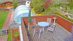Une terrasse en bois avec une table ronde et des chaises, un parasol vert et une structure de serre en verre.