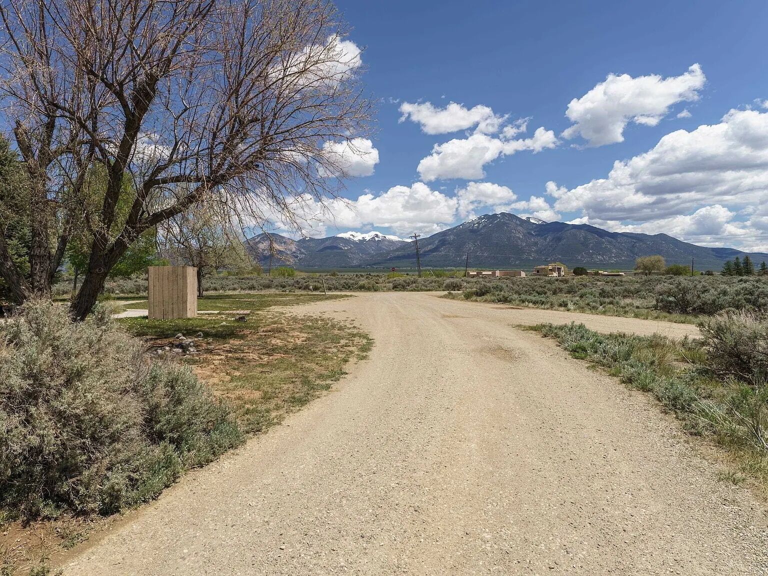 A gravel road leading to a wooden structure, surrounded by sparse vegetation and mountains in the background.