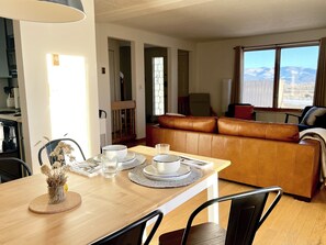A dining area with a table set for two, a brown leather sofa, and a view of mountains through a window.