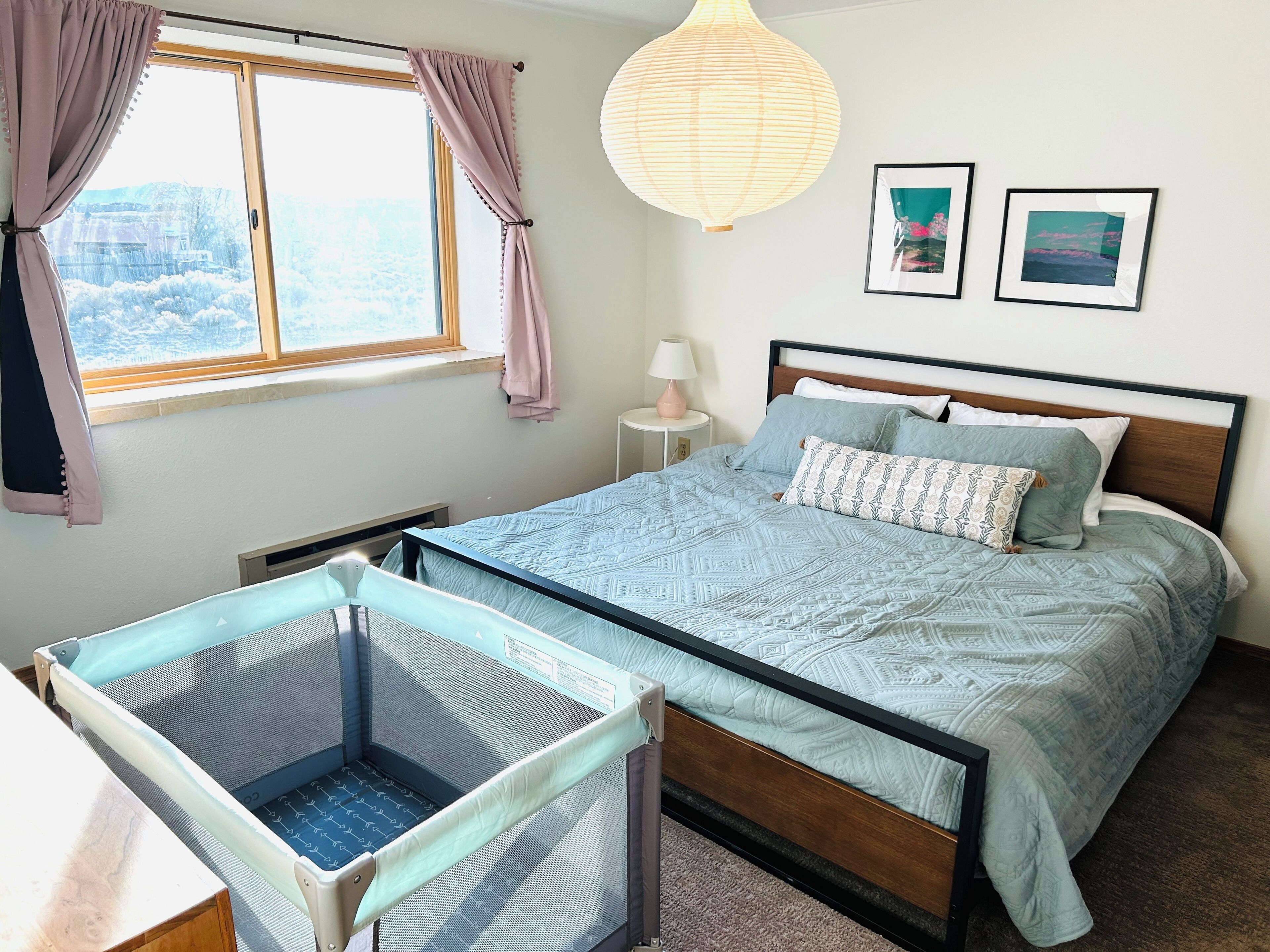 Quarto com cama, berço, janela com cortinas, abajur e duas fotos emolduradas na parede.