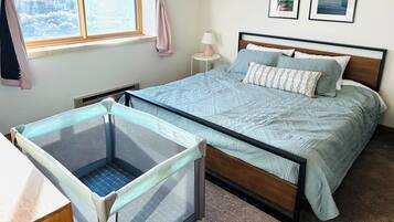 Quarto com cama, berço, janela com cortinas, abajur e duas fotos emolduradas na parede.