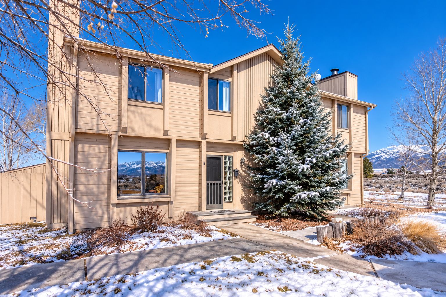 A two-story building with a large evergreen tree in front, a clear sky, and mountains in the background.