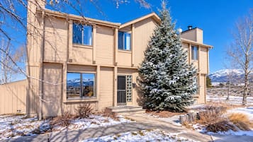 A two-story building with a large evergreen tree in front, a clear sky, and mountains in the background.
