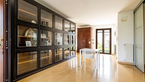 Una habitación amplia con un mueble grande de madera oscura y puertas de cristal, una mesa blanca con sillas, y vistas al exterior a través de un ventanal.