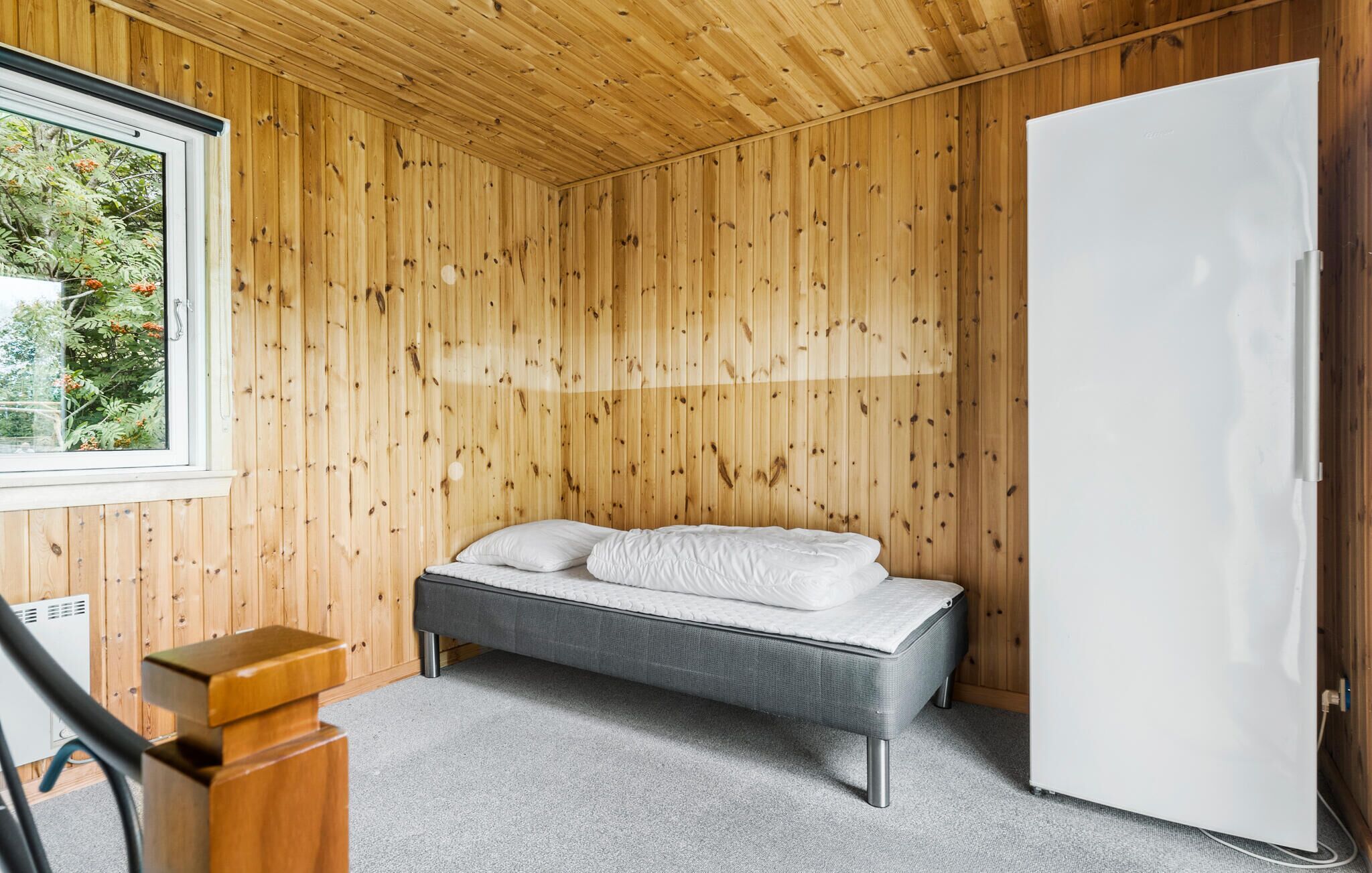 A small, cozy room with a wooden paneled wall, a bed with white bedding, a white refrigerator, and a window with a view of greenery.