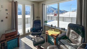 A cozy room with a view of a snowy landscape, featuring a large window, a chair, a small table, and a chessboard.