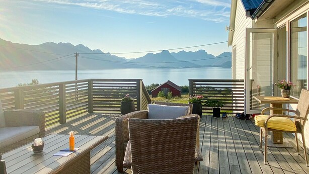 A wooden deck with wicker furniture, a small table, and a chair. A red building is visible in the background.