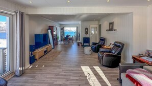 A spacious living room with a large TV, leather chairs, a fireplace, and a view of the sea.