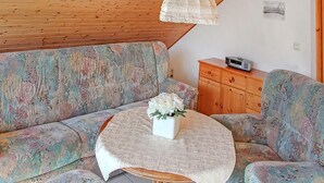 A cozy room with a wooden ceiling, a floral sofa, a small round table with a white vase, and a wooden cabinet with a radio. - Amazing apartment in Klink with sauna (Klink)