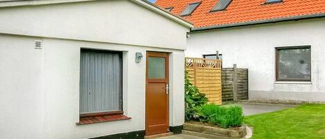 A single-story house with a white exterior, a brown door, and a window with a shutter. There is a small garden area with grass and plants.