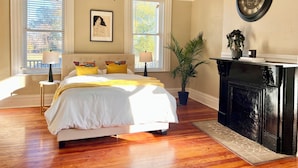 Quarto com cama grande, duas mesas de cabeceira com luminárias, lareira e uma janela ampla com persianas.