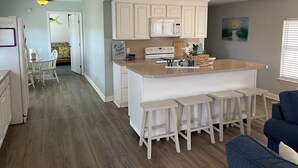 A modern kitchen with white cabinets, a granite countertop, and a built-in oven. There are three white bar stools and a blue couch in the dining area. - OCEANSIDE BEACH HOUSE-3 BED/2 BATH. GOLF-CART INCLUDED, 100 YARDS FROM BEACH (Myrtle Beach)