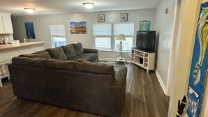 A modern living room with a sectional sofa, a flat-screen TV, and a dining area with a white table and chairs. - OCEANSIDE BEACH HOUSE-3 BED/2 BATH. GOLF-CART INCLUDED, 100 YARDS FROM BEACH (Myrtle Beach)