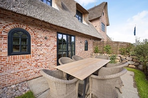 Une maison en briques avec un toit de chaume, une table en bois, des chaises en osier et un mur en pierre.