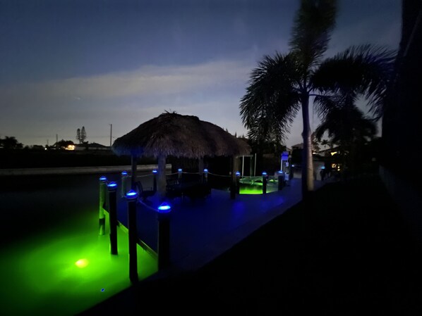A thatched-roof structure with a pool area, surrounded by palm trees and illuminated by blue and green lights. - Gulf-Access Luxury – Heated Pool, Hot Tub, Tiki Hut, Pool Table and more! (Cape Coral)