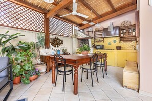 Uma sala de jantar com uma mesa e cadeiras de madeira, cercada por plantas em vasos, e uma cozinha com armários amarelos e um fogão.