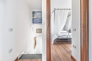 Un couloir mène à une chambre à coucher où se trouvent un lit, un rideau blanc et un tableau accroché au mur.