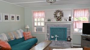 A living room with a teal sofa, a fireplace, a television, and framed pictures on the wall. - Cape Cottage with 5 beds, large deck, and less than 10 minute walk to the beach! (Eastham)