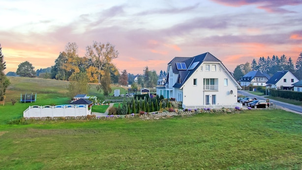 Een woonwijk met een opvallend wit huis met een balkon, omringd door weelderige begroeiing en een heldere lucht.