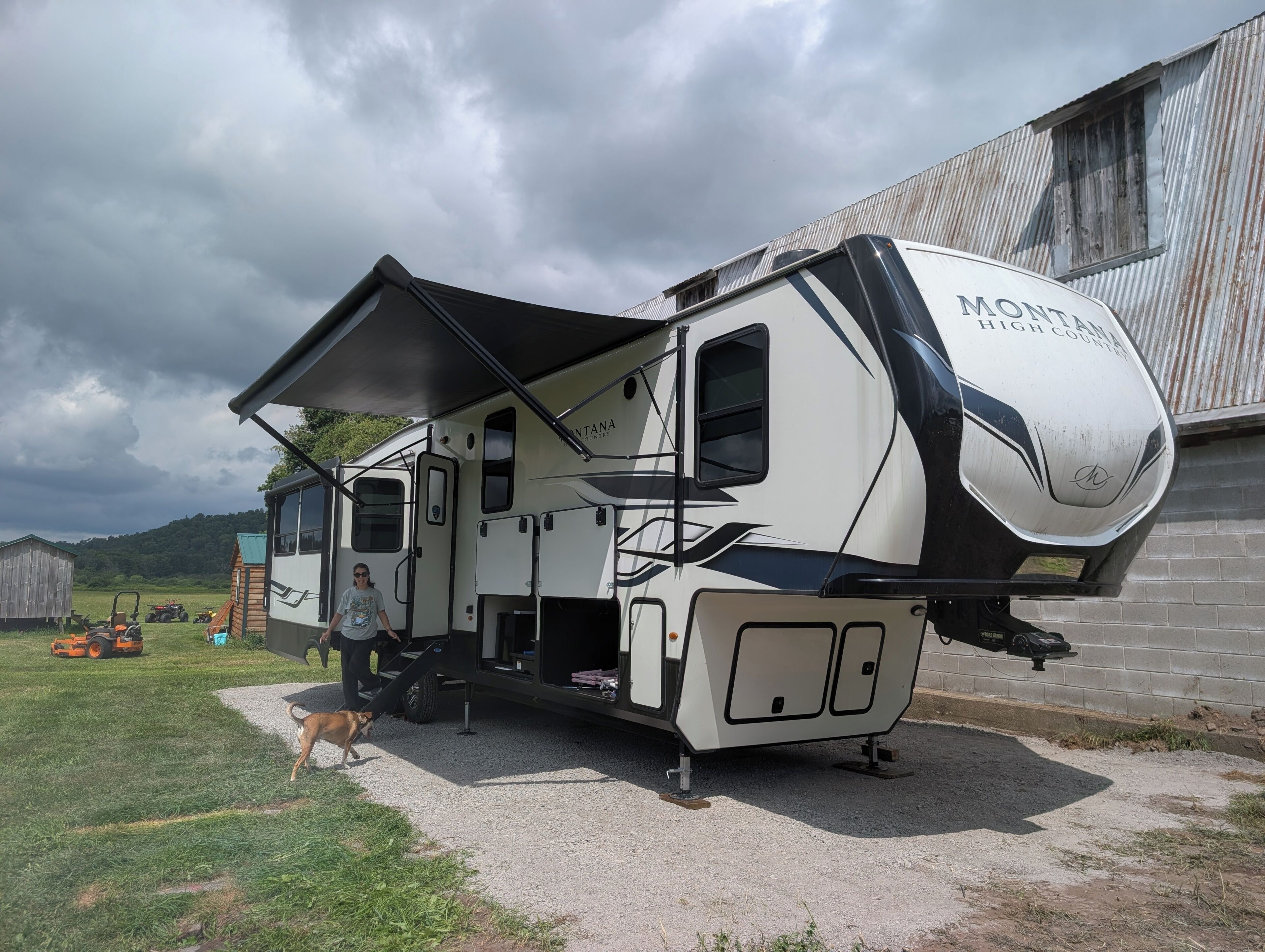 A recreational vehicle parked on a gravel surface with a covered porch, a dog nearby, and a barn in the background.