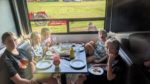A group of people seated at a table inside a vehicle with a view of a grassy field and a red tractor outside.