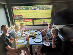 A group of people seated at a table inside a vehicle with a view of a grassy field and a red tractor outside.