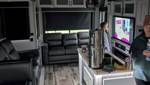 A compact RV interior with a kitchenette, seating area, and a television displaying a show.