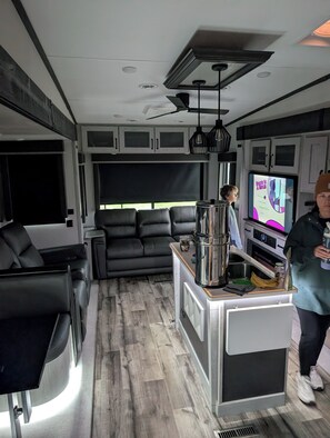 A compact RV interior with a kitchenette, seating area, and a television displaying a show.
