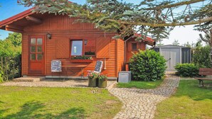 A wooden cabin with a red door, a window with a red awning, a small porch with a table and chairs, and a garden with a hedge and a shed. - Stunning home in Kummerow with sauna (Kummerow)