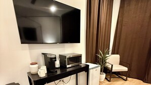 A modern living room with a flat-screen TV, a coffee maker, a microwave, a small refrigerator, a trash bin, a black rug, and a potted plant. - Luxurious King Suite in Gilbert. Kids are Welcome! (Gilbet)