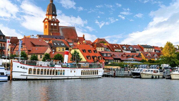 A riverfront with a historic building featuring a clock tower, a white boat docked at the pier, and colorful houses with red roofs.