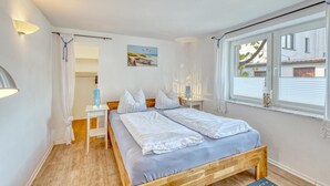 A bedroom with a wooden bed, a bedside table, a nightstand, a window with blinds, and a wall-mounted lamp.