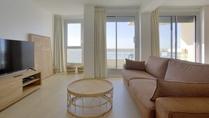 A modern living room with a flat-screen TV, a brown sofa, a wicker coffee table, and large windows with curtains. - Appartement Victoire, vue mer - Welkeys (Biarritz)