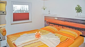 Ein ordentlich bezogenes Bett mit gestreifter Bettwäsche, ein Fenster mit rotem Rollo und eine kleine Pflanze auf einem Nachttisch.