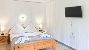 Une chambre à coucher avec un lit en bois, du linge de lit blanc, deux lampes de chevet, un téléviseur à écran plat fixé au mur et une prise de courant.