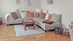 Een woonkamer met een hoekbank, een glazen salontafel, een kroonluchter en een spiegel aan de muur.