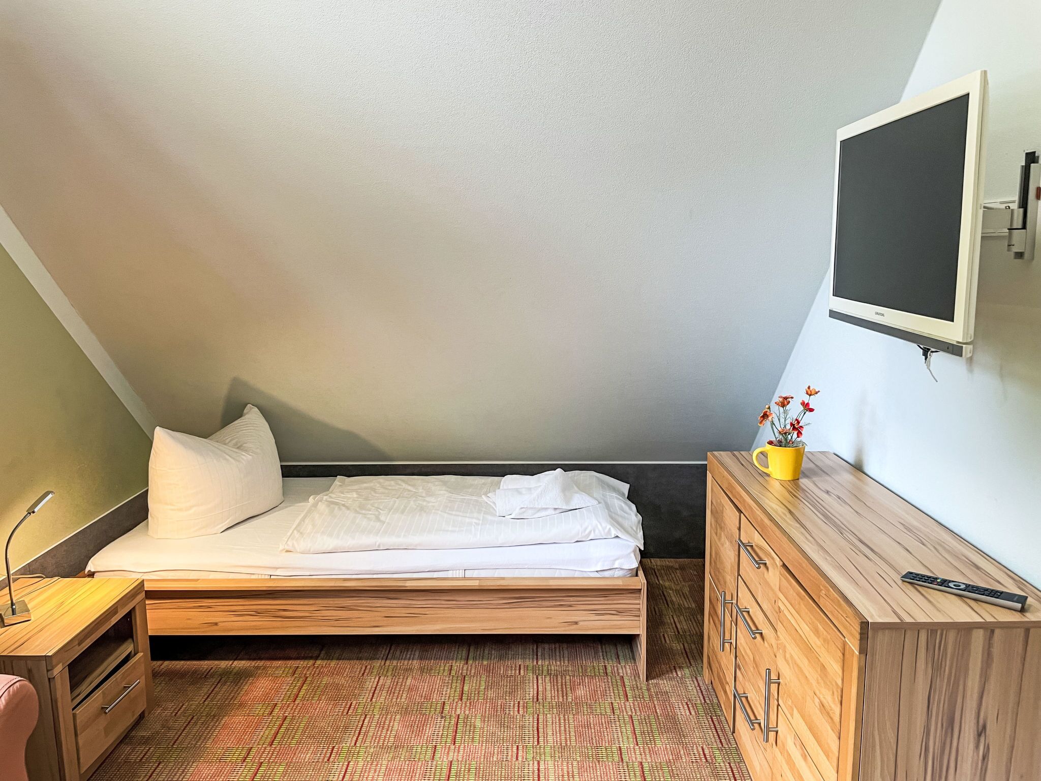 Une petite chambre à coucher, bien rangée, avec un lit, une commode en bois, une table de chevet et un téléviseur fixé au mur.