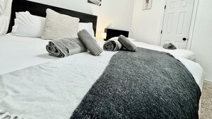 Una cama bien hecha con toallas enrolladas y una manta, un cabecero, una mesita de noche con una lámpara y una puerta en una habitación sencilla.
