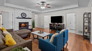 Una sala de estar con chimenea, ventilador de techo, televisor y estantería.