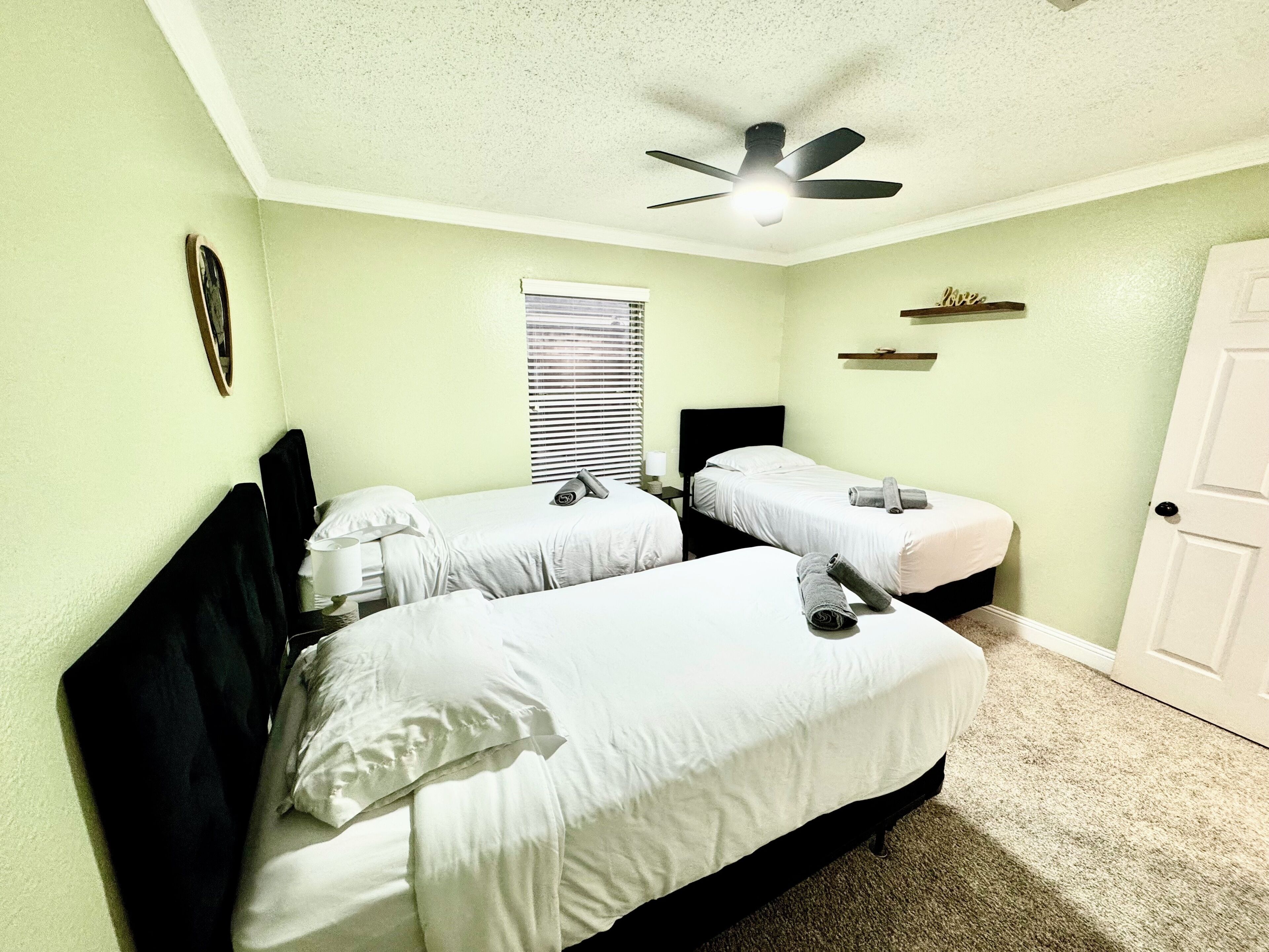 A room with two beds, a ceiling fan, a window with blinds, and a door.