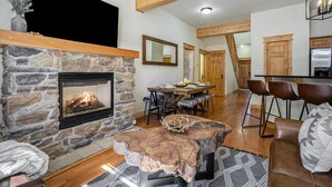 A cozy living room with a stone fireplace, a dining area with a wooden table and chairs, and a kitchen with wooden cabinets and a stainless steel refrigerator. - Estes Park Luxury Family Retreat- EV Charger/Hot Tub/Game Room/Outdoor Fire Pit (Estes Park)