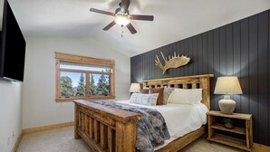 A bedroom with a wooden bed, a ceiling fan, a mounted deer head, a window with a view of trees, and a small bedside table with a lamp and a plant. - Estes Park Luxury Family Retreat- EV Charger/Hot Tub/Game Room/Outdoor Fire Pit (Estes Park)