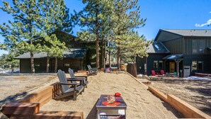 A wooden deck with Adirondack chairs, a table with a red cushion, and a building with a sloped roof in the background. - Estes Park Luxury Family Retreat- EV Charger/Hot Tub/Game Room/Outdoor Fire Pit (Estes Park)