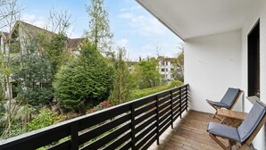 A balcony with wooden flooring, two lounge chairs, and a view of surrounding buildings and greenery.
