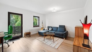 A modern living room with a blue sofa, a small coffee table, a green office chair, a radiator, and a window with a view of trees.
