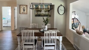 Un comedor con una mesa de madera, sillas blancas, un tapete estampado, una lámpara colgante y un reloj en la pared.