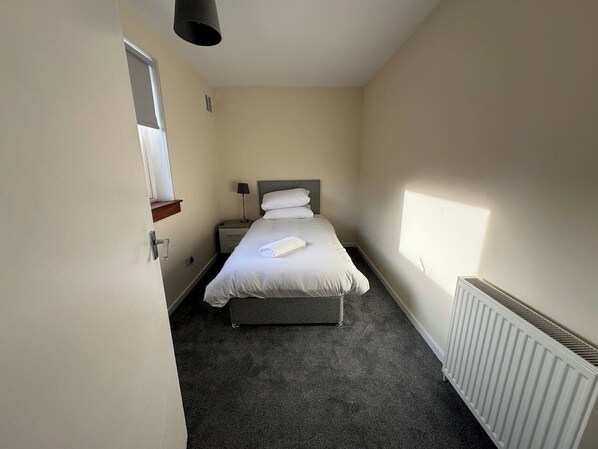 Un dormitorio pequeño y ordenado con una cama individual, una mesita de noche, una lámpara y un radiador.