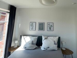 A modern hotel room with a large bed, two bedside tables, and a window with curtains. There are three framed pictures on the wall above the bed. - Zweite Heimat - Hochwertige Neubauwohnung Direkt am Wasser! (Krummhörn)