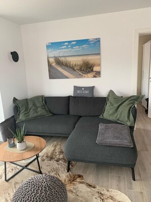 A modern living room with a sectional sofa, a small round table with a plant, and a large painting of a beach scene. - Zweite Heimat - Hochwertige Neubauwohnung Direkt am Wasser! (Krummhörn)
