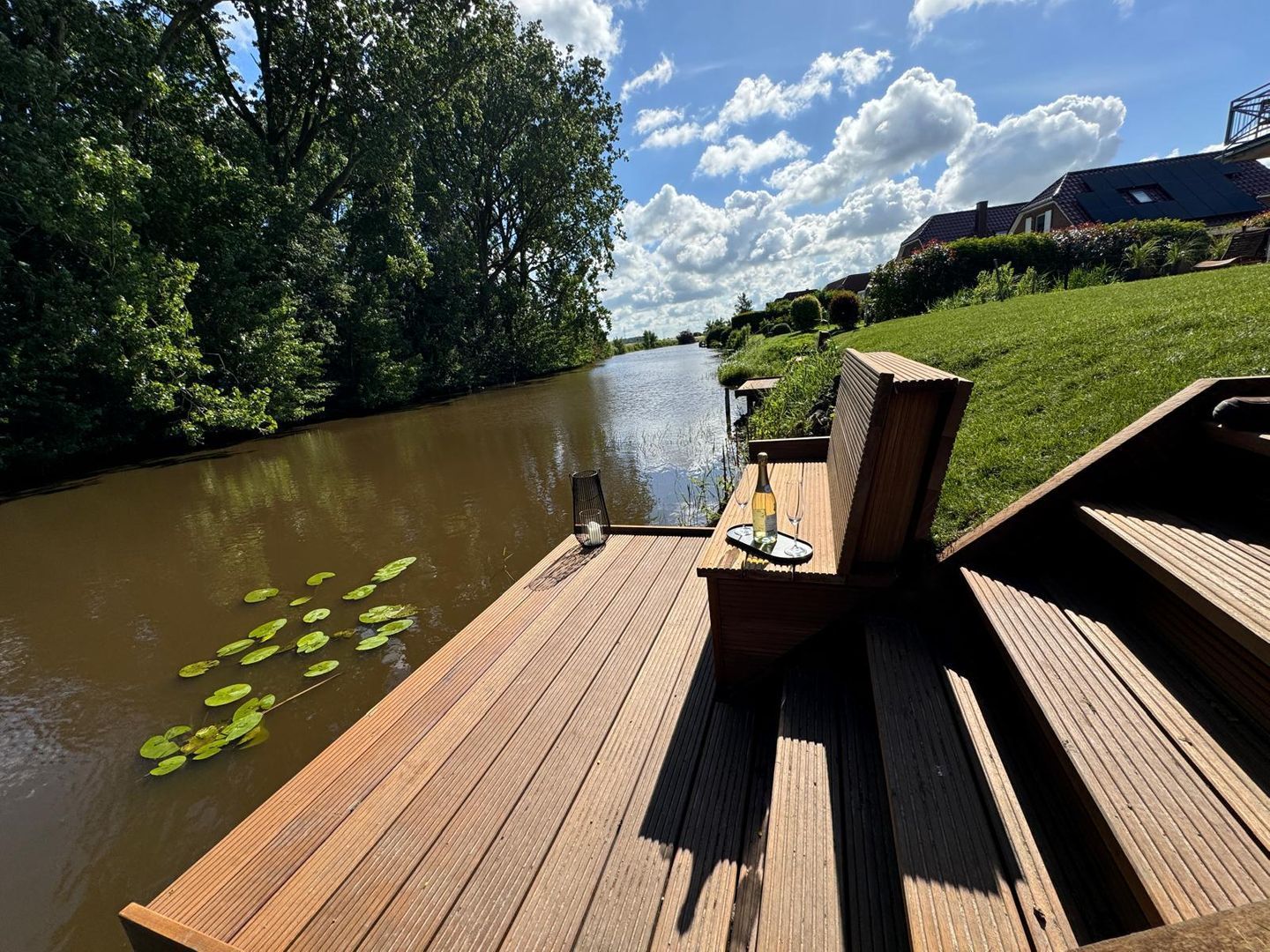 A wooden deck with a table and chairs overlooking a river, surrounded by greenery and a clear sky.